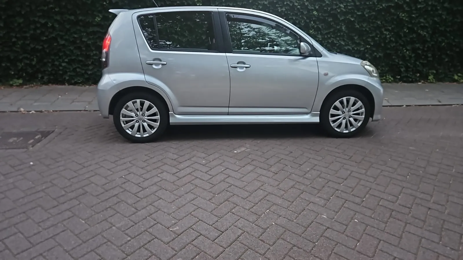 Zilvergrijze Daihatsu Sirion Sport voor een heg